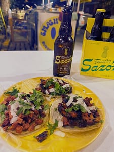 three tacos on a plate with a bottle of sazon tepache